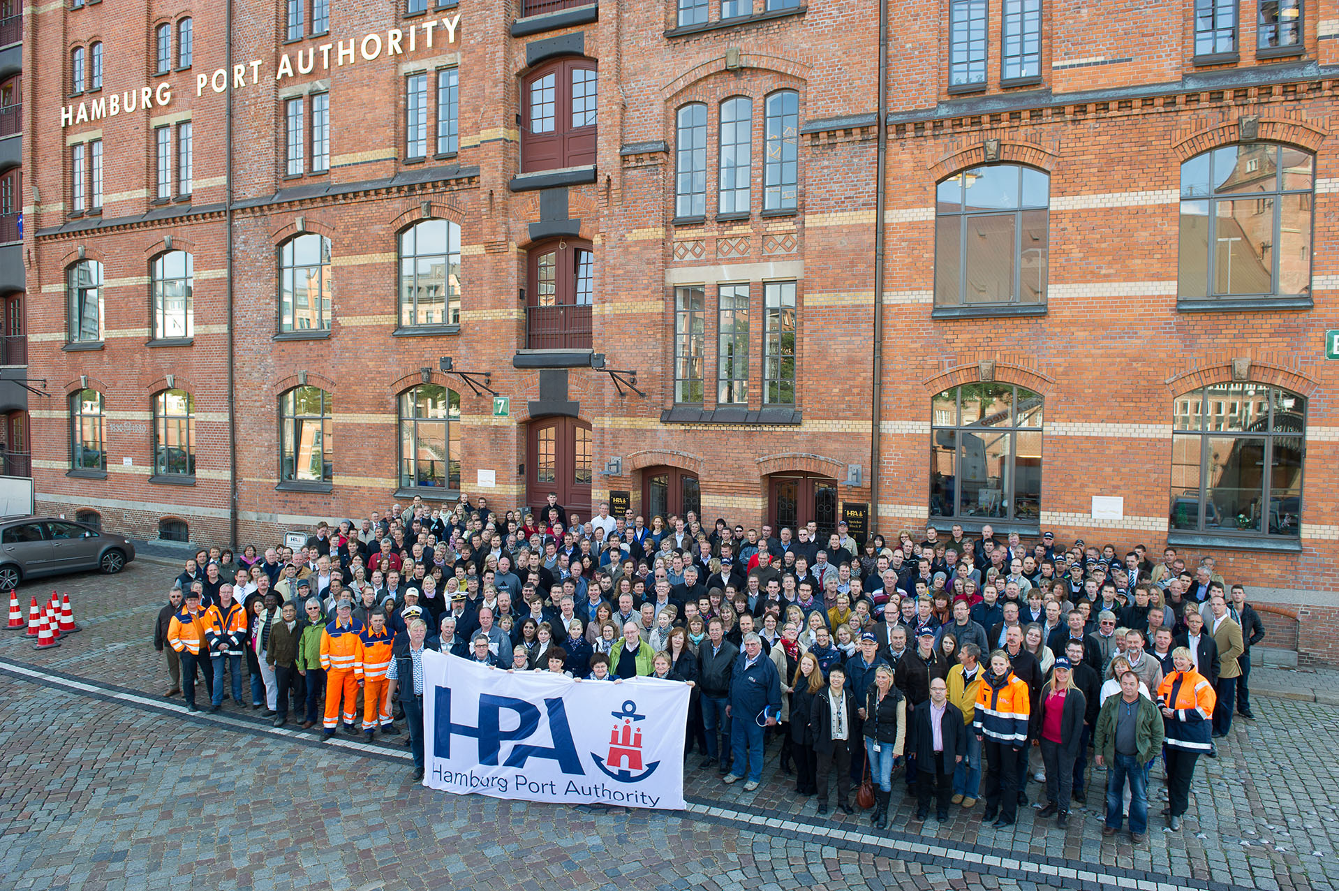 Eine Gruppe von Mitarbeitenden der Hamburg Port Authority (HPA) steht vor der Unternehmenszentrale, einem historischen Backsteingebäude in der Hamburger Hafencity.